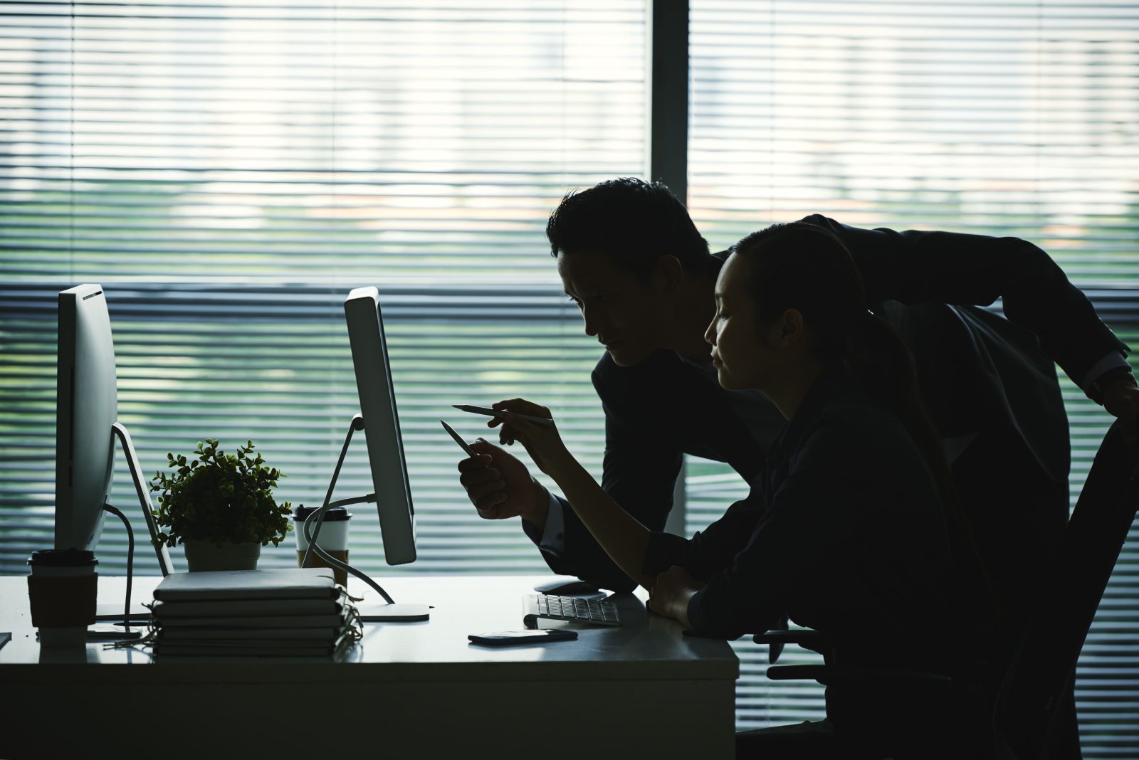dark-silhouettes-colleagues-pointing-computer-screen-office-against-window 1